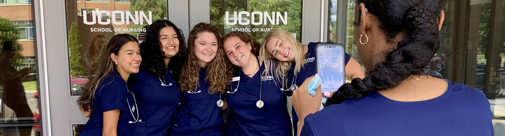 A group of women in scrubs pose in front of a building's doors while another woman takes their photo on a cellphone